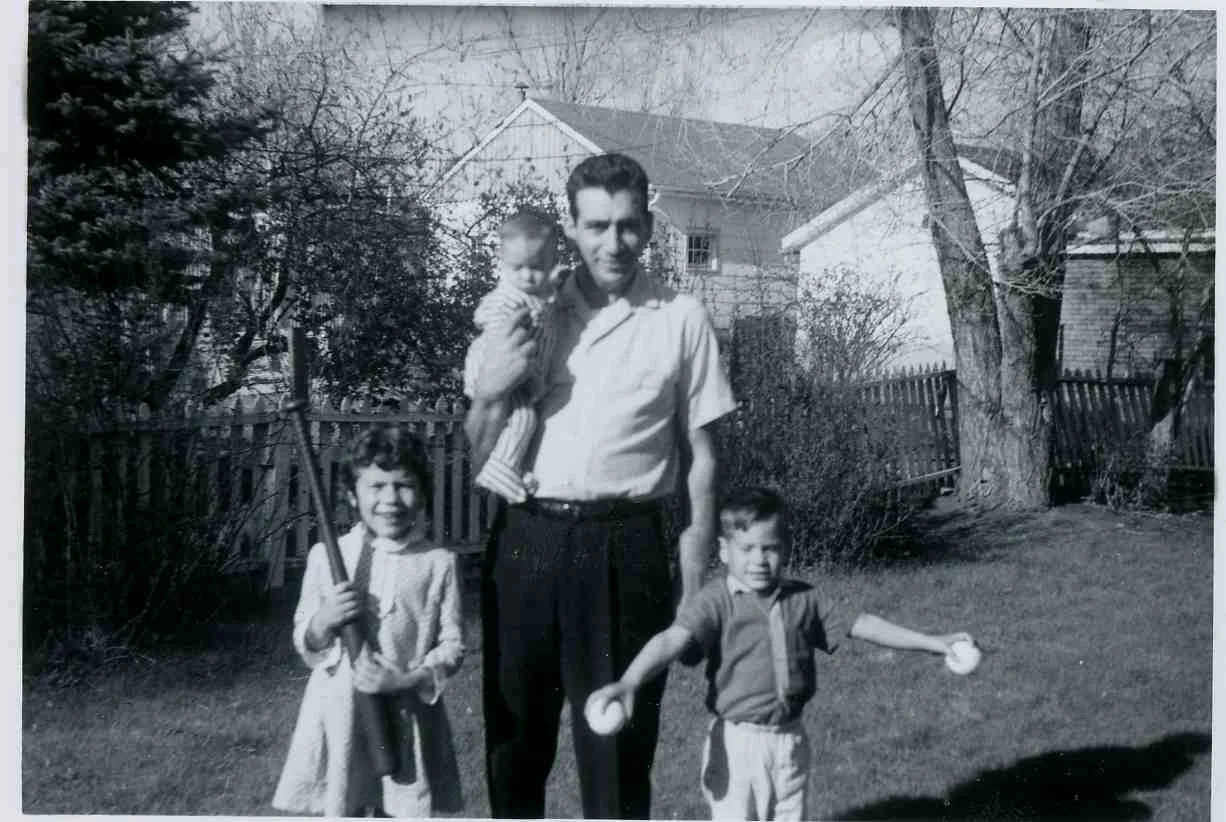 A father holding a baby, standing in a backyard with two young children, in 1966.