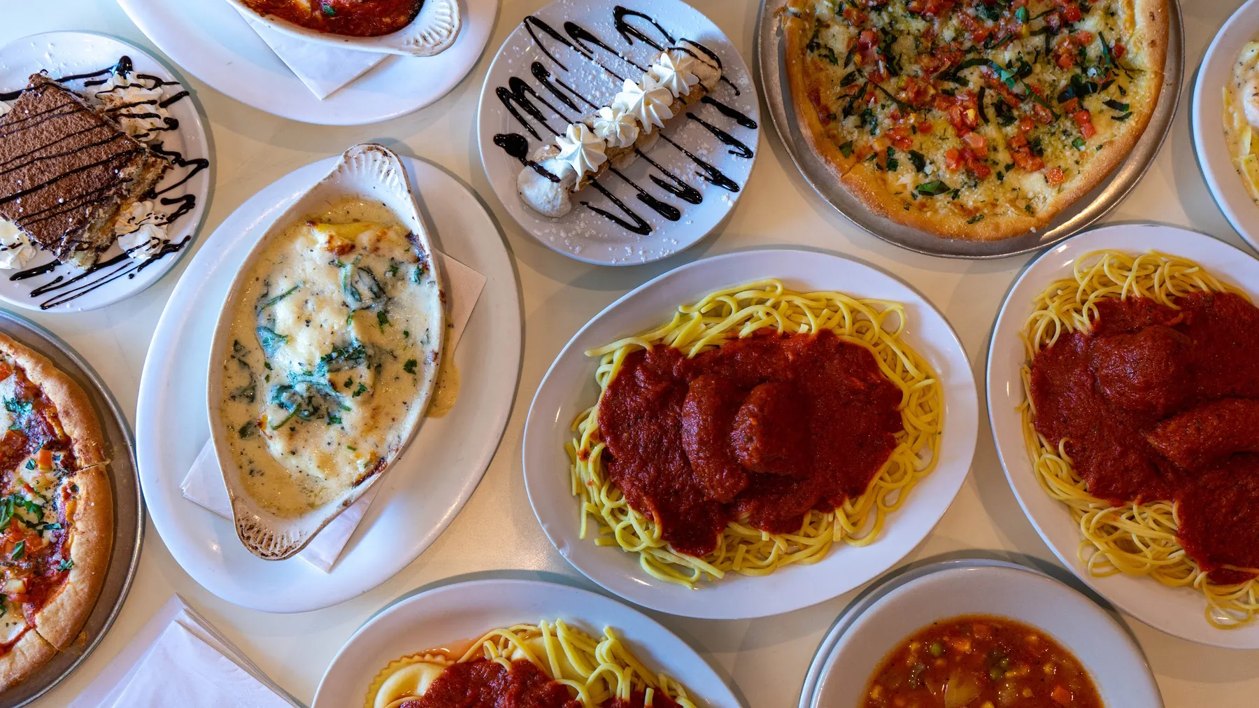 An overhead spread of Romano's dishes: tiramisu, spinach artichoke dip, cannoli, brick oven pizza, spaghetti with meat sauce, ravioli and minestrone.